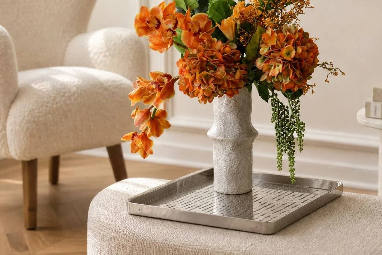 Levande orange blommor i en texturerad vit vas på en silverbricka, placerad på en ljus ottoman i ett modernt vardagsrum.