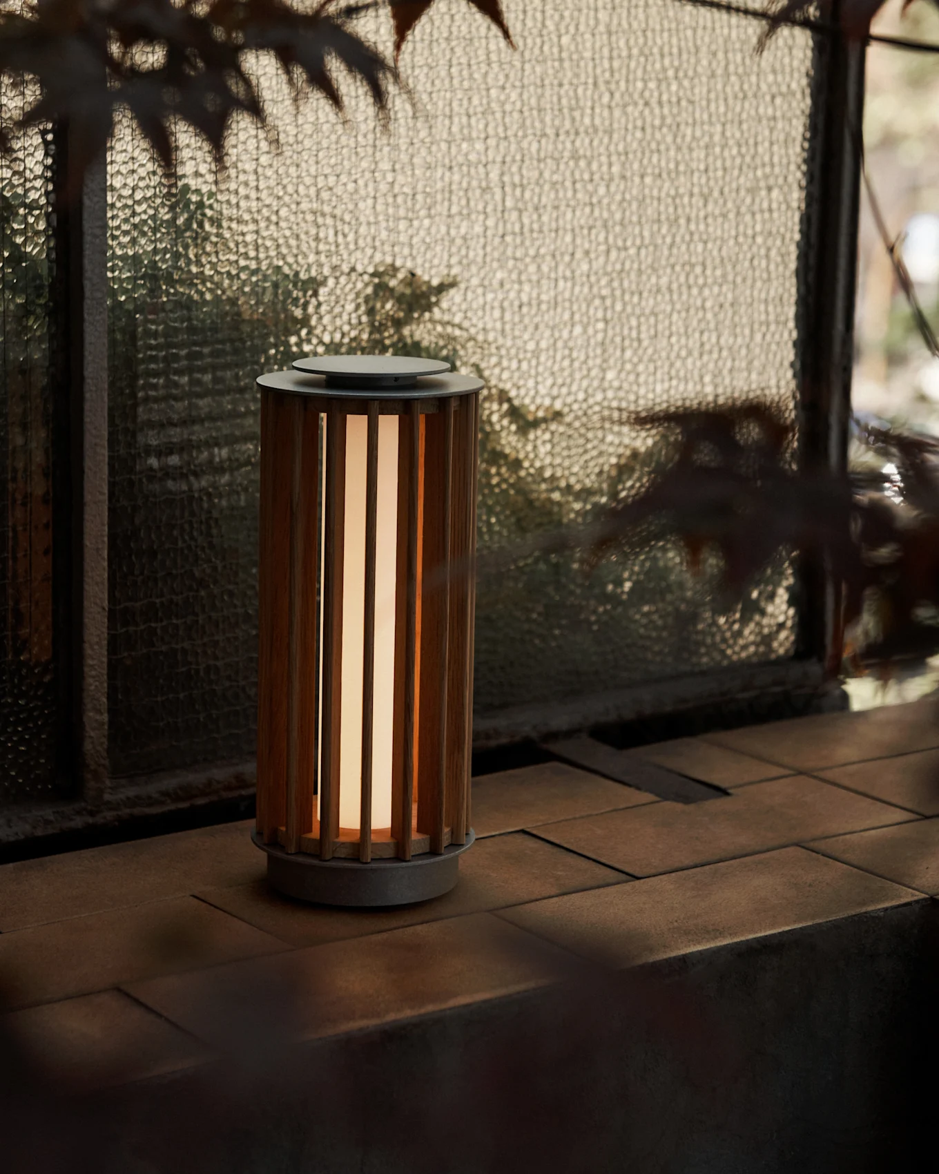 En cylindrisk bärbar lampa med en ribbad träkropp och varmt sken, på en kaklad yta vid ett texturerat glasfönster.