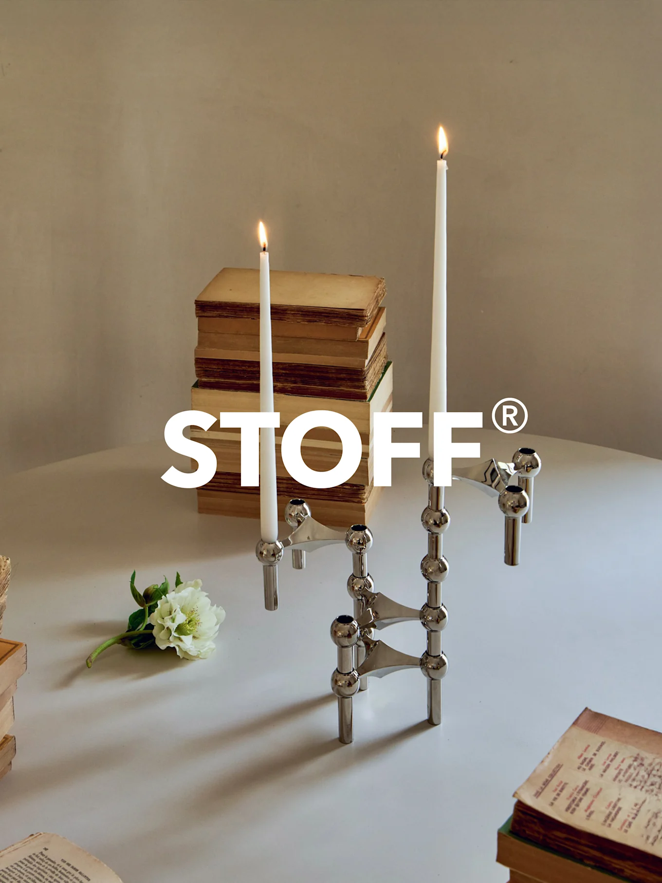 Polerad krom modulär kandelaber med två tända ljus, omgiven av gamla böcker och en vit blomma på ett bord. STOFF® logotyp.