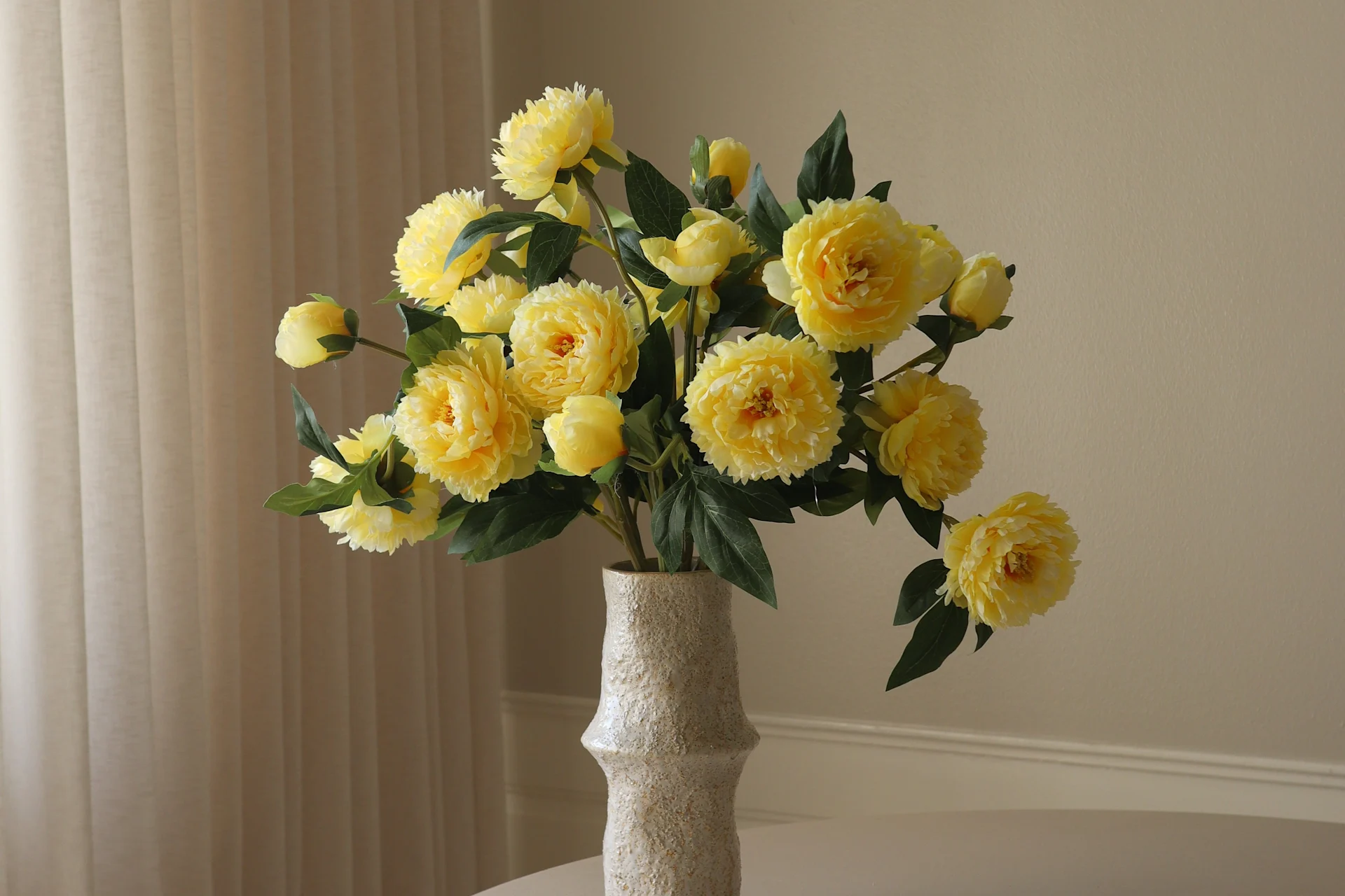 En ljus, texturerad vas innehåller en levande bukett med gula pionliknande blommor och gröna blad mot en ljus bakgrund.