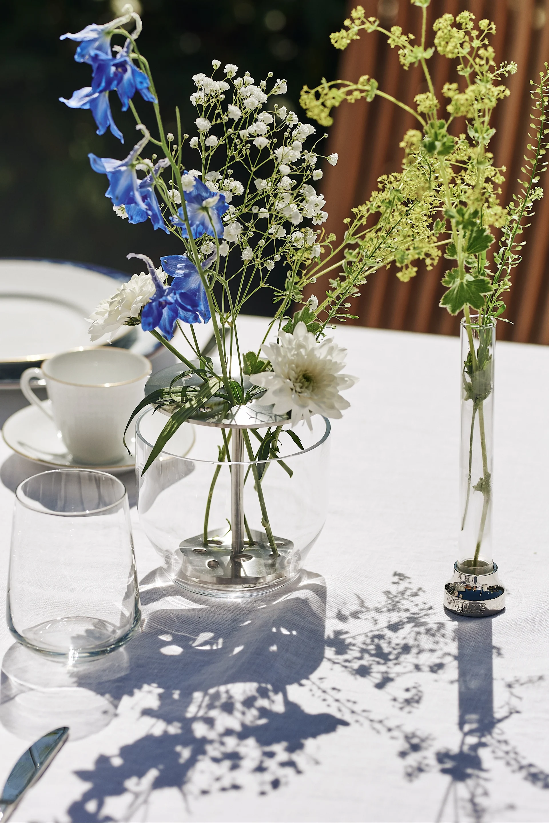 En blåvit och klassisk midsommardukning står i trädgården med blåvitt porslin, vaser och detaljer i glas och rostfritt stål, här Ikebana vas i glas och stål med blåa och vita blommor i, placerad på vit linneduk.