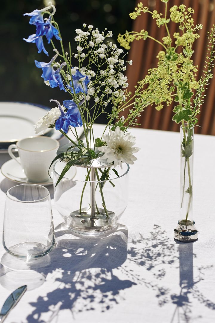En blåvit och klassisk midsommardukning står i trädgården med blåvitt porslin, vaser och detaljer i glas och rostfritt stål, här Ikebana vas i glas och stål med blåa och vita blommor i, placerad på vit linneduk.