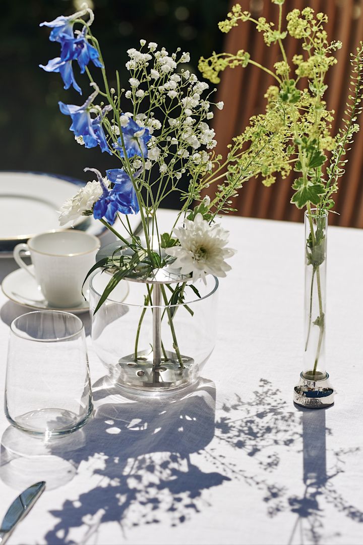 En blåvit och klassisk midsommardukning står i trädgården med blåvitt porslin, vaser och detaljer i glas och rostfritt stål, här Ikebana vas i glas och stål med blåa och vita blommor i, placerad på vit linneduk.