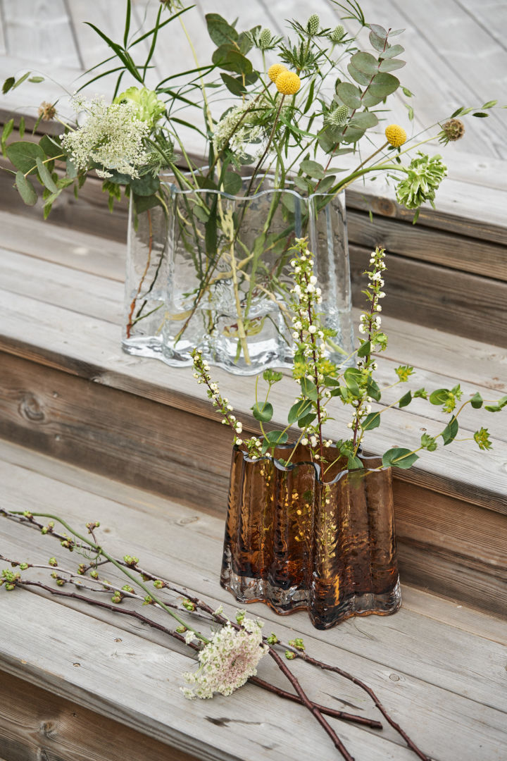 Gry vasen i färgerna clear och cognac står på ett altandäck i trä med vilda vårblommor i sig.