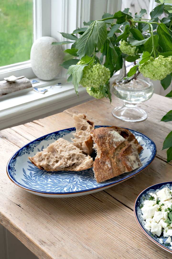 Skapa en midsommardukning i klassiskt blåvitt med hjälp av Ostindia Floris-serien från Rörstrand, här ovalt serveringsfat fyllt med bröd, placerat på rustikt träbord vid fönster.