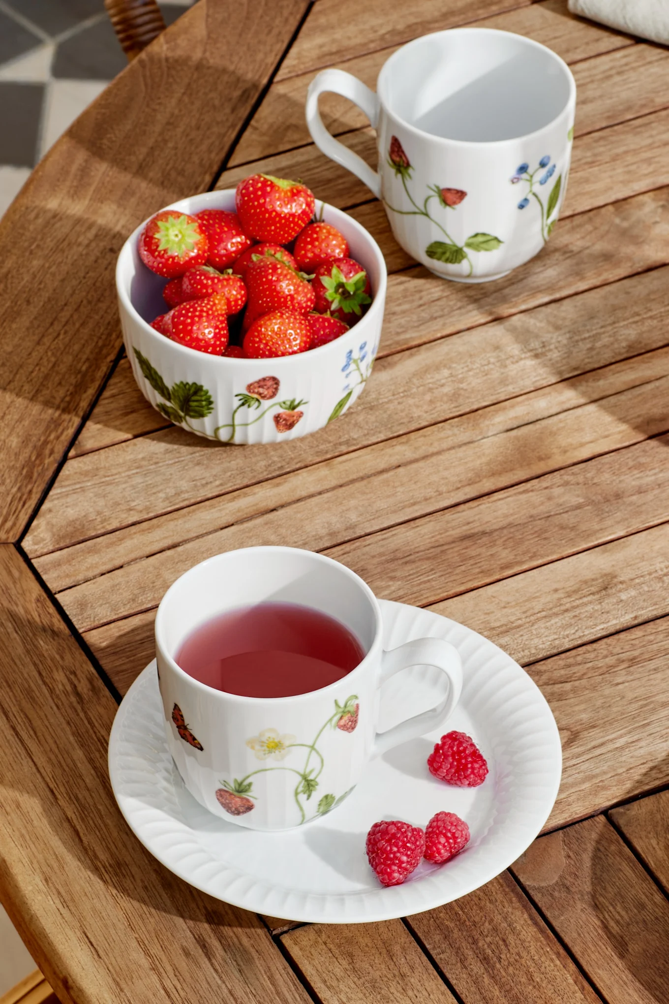 En vit mugg med rosa te och ett fat med hallon, en skål jordgubbar och en tom mugg står på ett träribbord i solljus.
