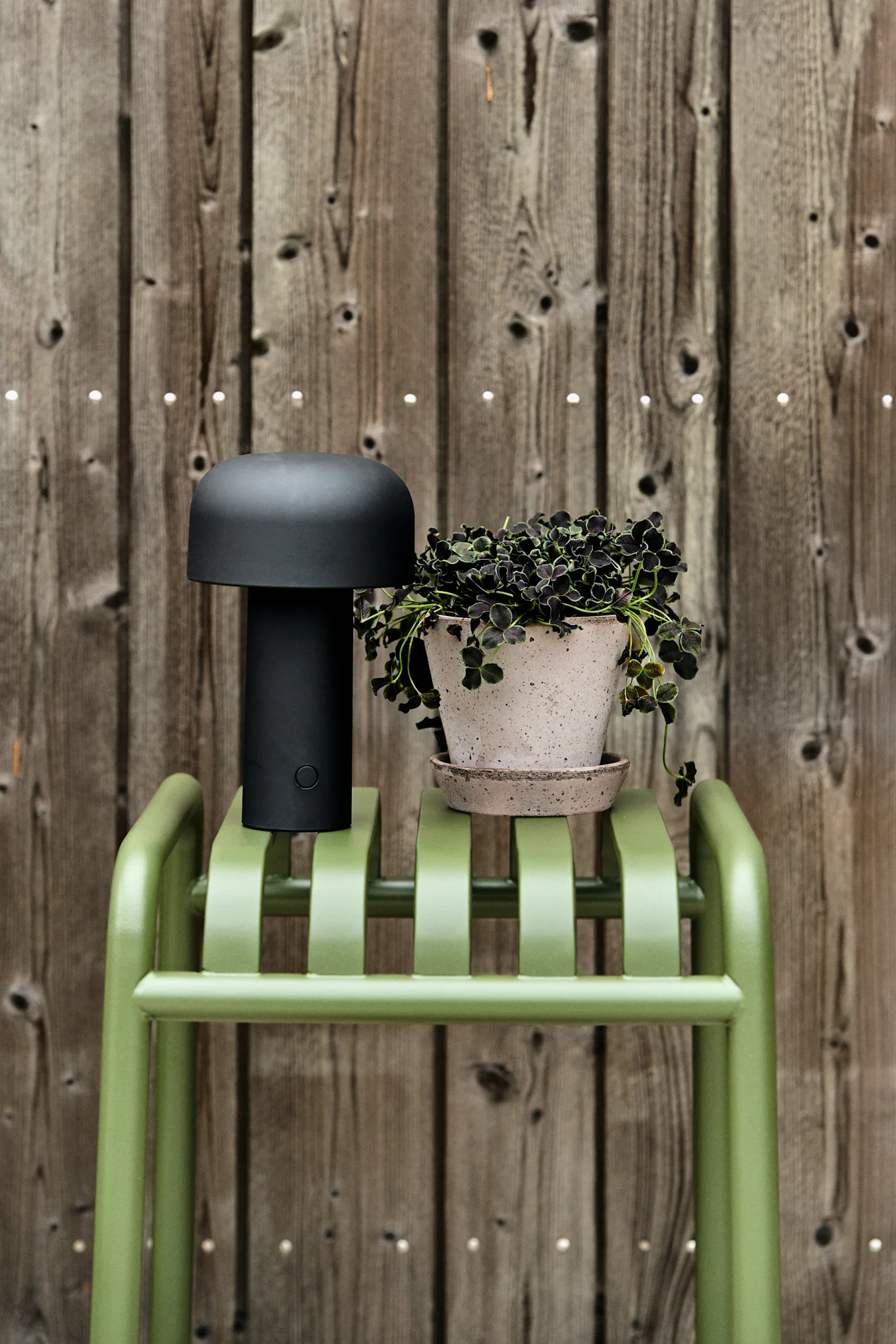 En svart svampformad lampa och en krukväxt med mörka blad står på en grön ribbad bänk mot ett trästaket. Inspiration till din balkong 2026.