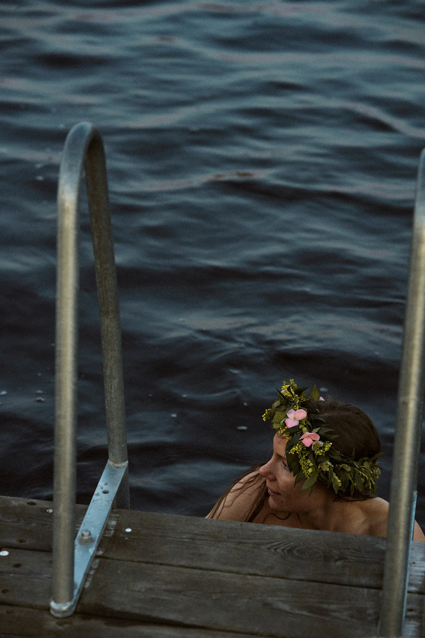 Kvinna i mörkt vatten med blomsterkrans, bredvid en träbrygga med metallräcken.
