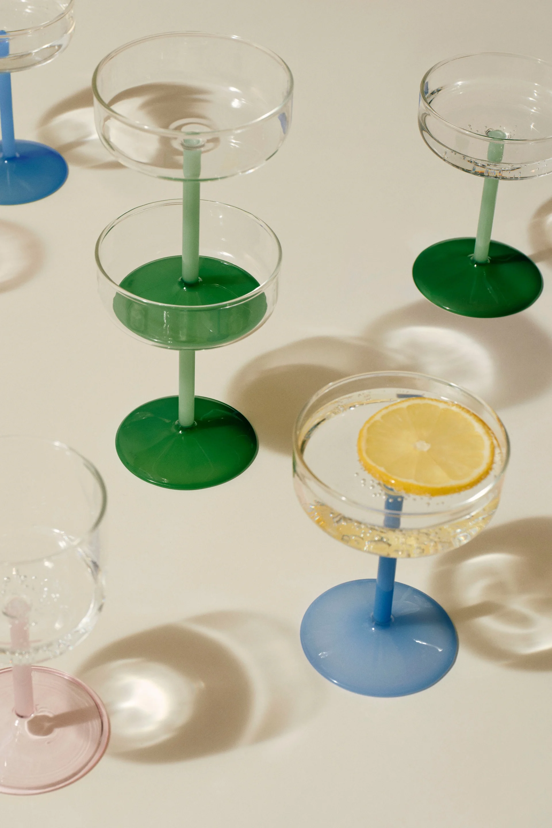 Sortiment av coupeglas med färgglada stjälkar och fötter. Ett glas innehåller en citronskiva i kolsyrat vatten.