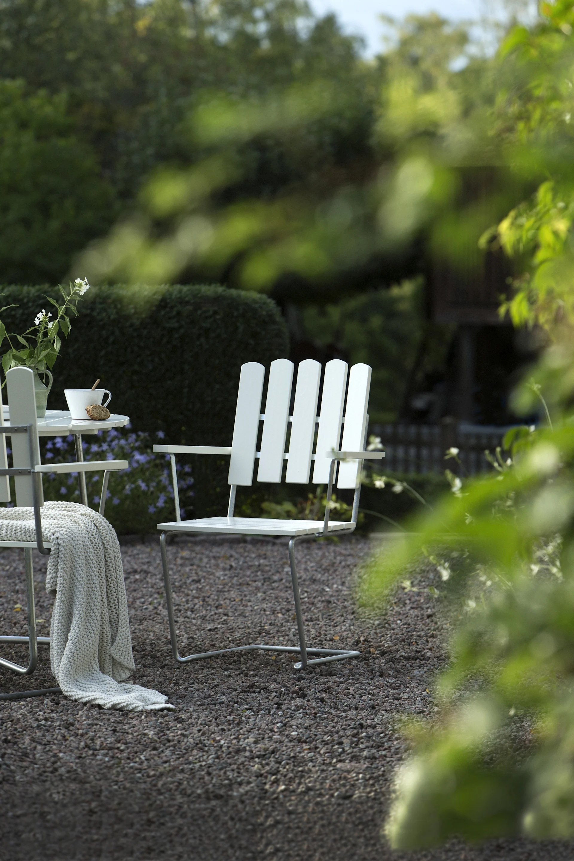 En vit trädgårdsstol och bord på grus, med blommor, en mugg och en filt, placerade mot en grön trädgård.
