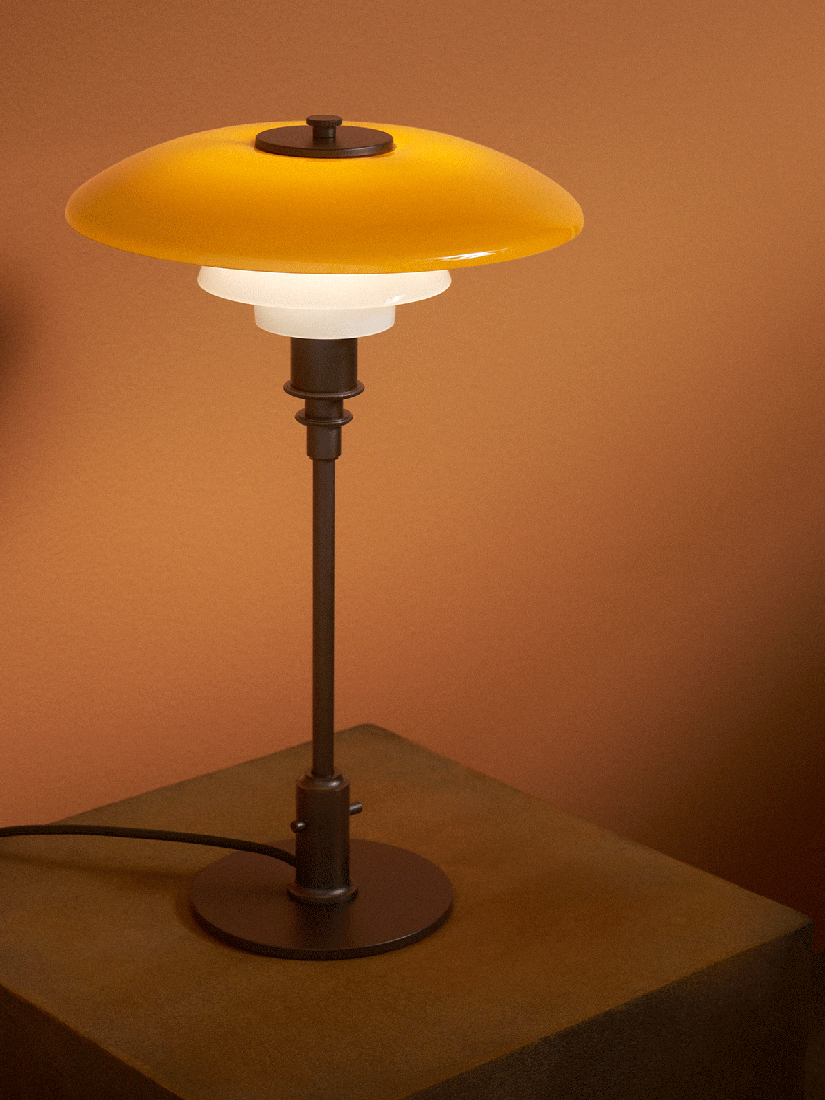 En modern bordslampa med gul övre skärm och två vita skärmar står på en texturerad träpiedestal mot en brun vägg.