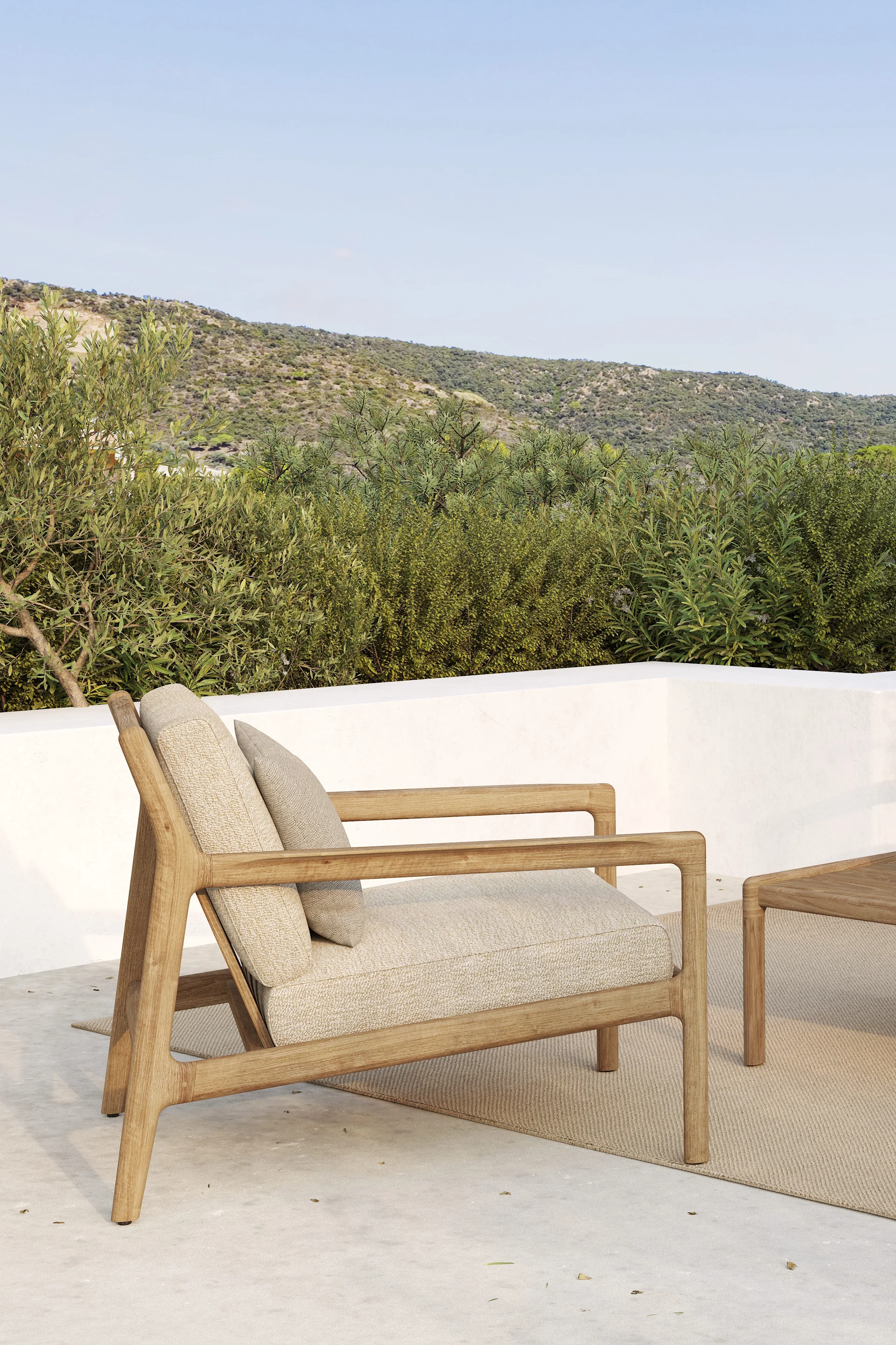 Utomhusfåtölj i trä med ljusa dynor på en uteplats, mot gröna kullar och en blå himmel.