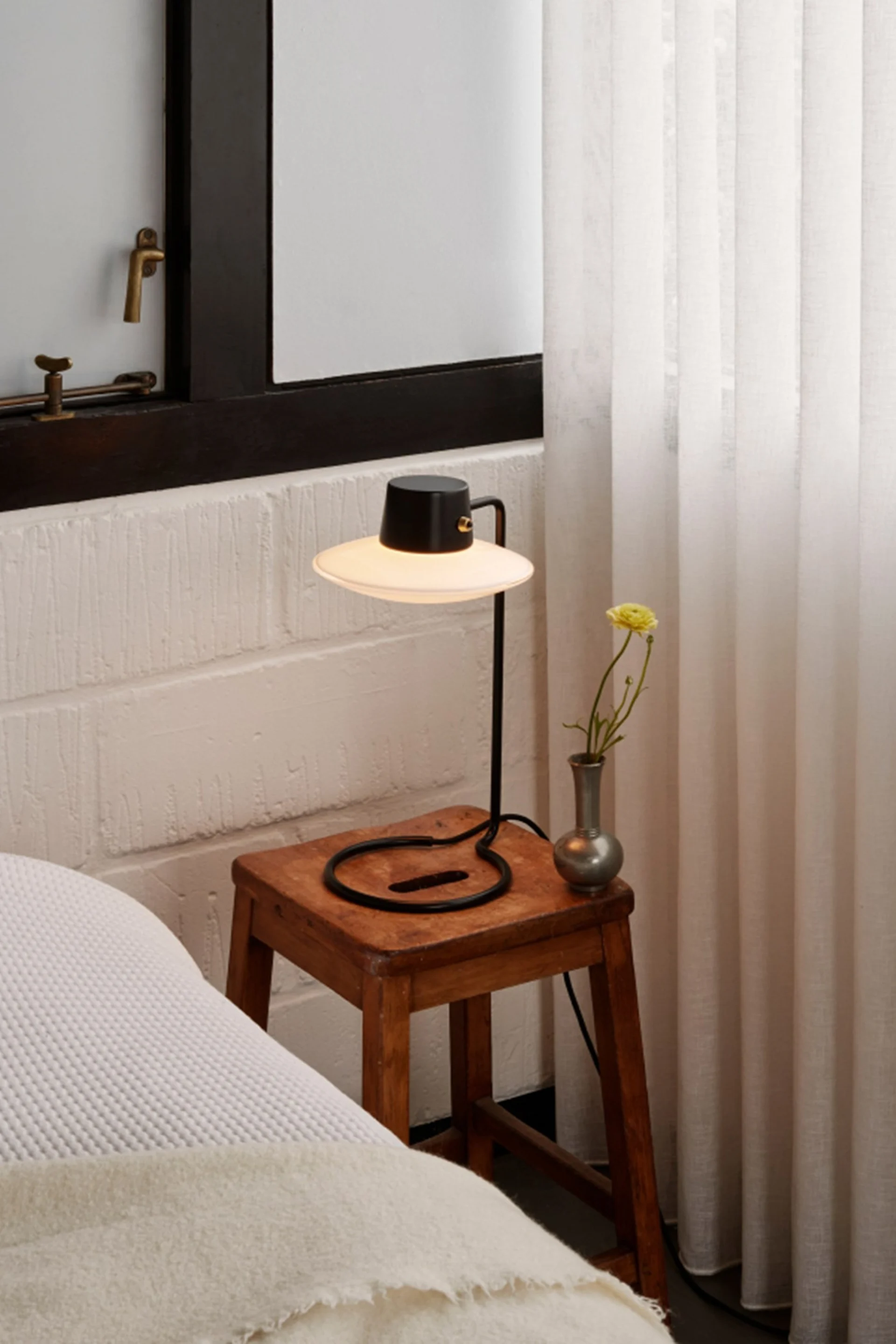 Svart bordslampa med bred skärm på rustik träpall, vas med gul blomma, bredvid säng och vita gardiner.