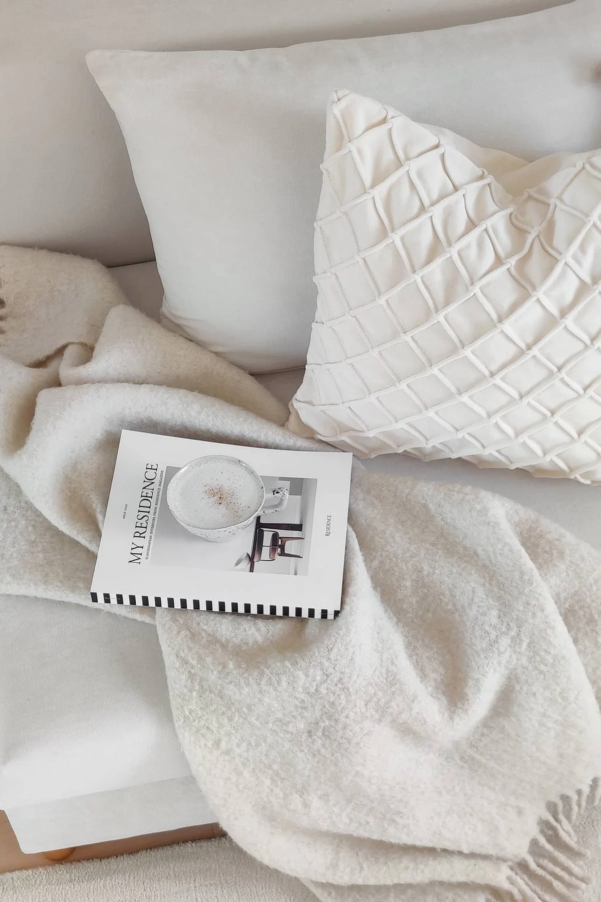 Alla hjärtans dag presenter: På en vit soffa ligger två vita kuddar från Chhatwal & Jonsson tillsammans med Ralph-pläden från Klippan Yllefabrik, en bok och en kopp kaffe.