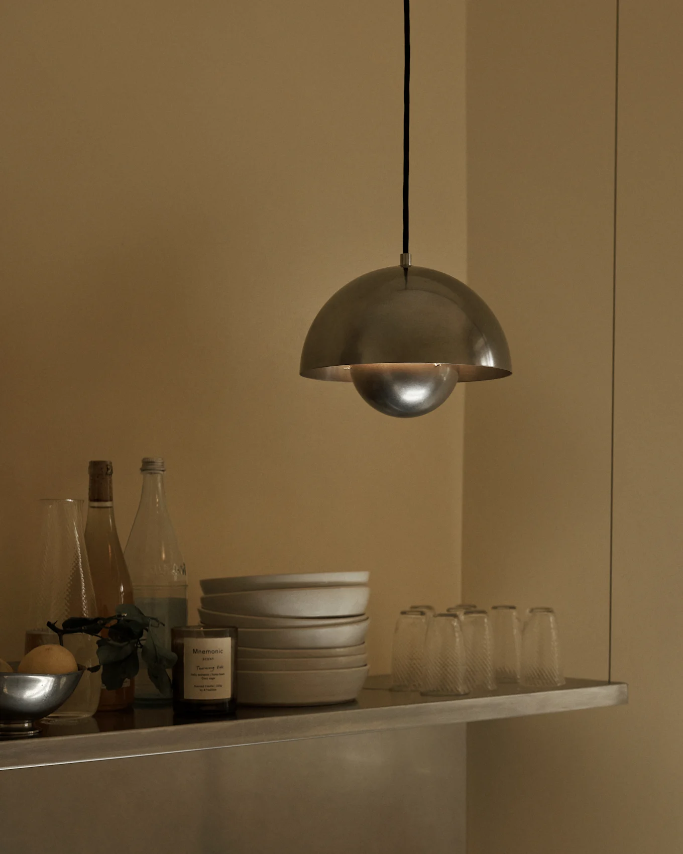 En taklampa med borstad metalldom hänger över en hylla med skålar, flaskor och glas.