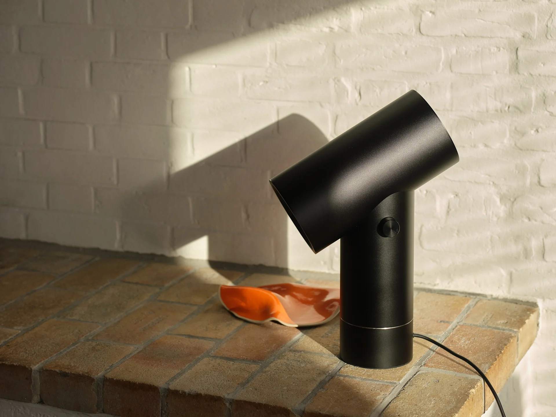 Svart cylindrisk lampa på en kaklad yta med en vit tegelvägg i solljus.
