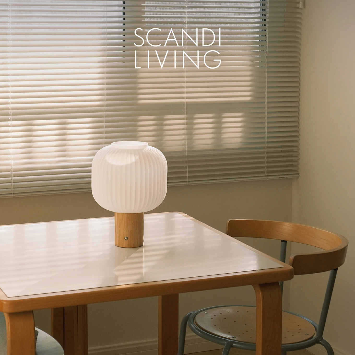 Vit räfflad bordslampa med träfot på ett vittoppat bord och en stol, framför fönsterpersienner. Text: SCANDI LIVING.