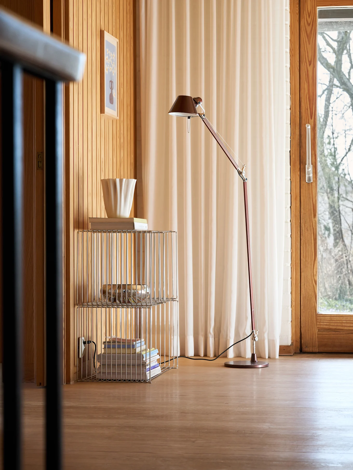 Ett mysigt rum med träväggar och golv, en röd golvlampa, en kromhylla med böcker och en räfflad vit vas, samt ett fönster med ljusa gardiner.