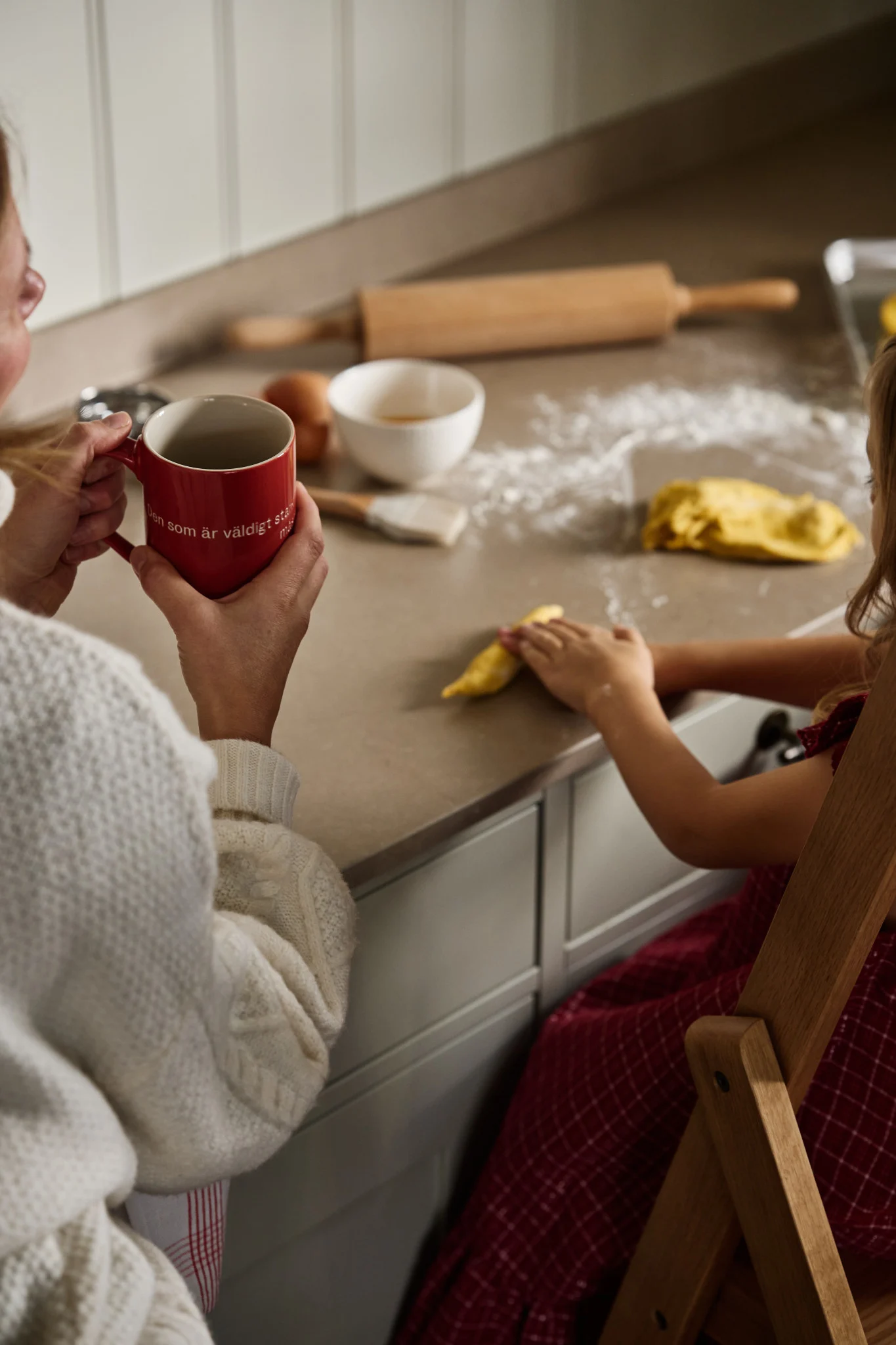 En vuxen och ett barn firar första advent med en mysig bakningsaktivitet. På köksbänken ligger saffransdeg, kavel och ägg, och den vuxna håller en röd mugg från Design House Stockholm.