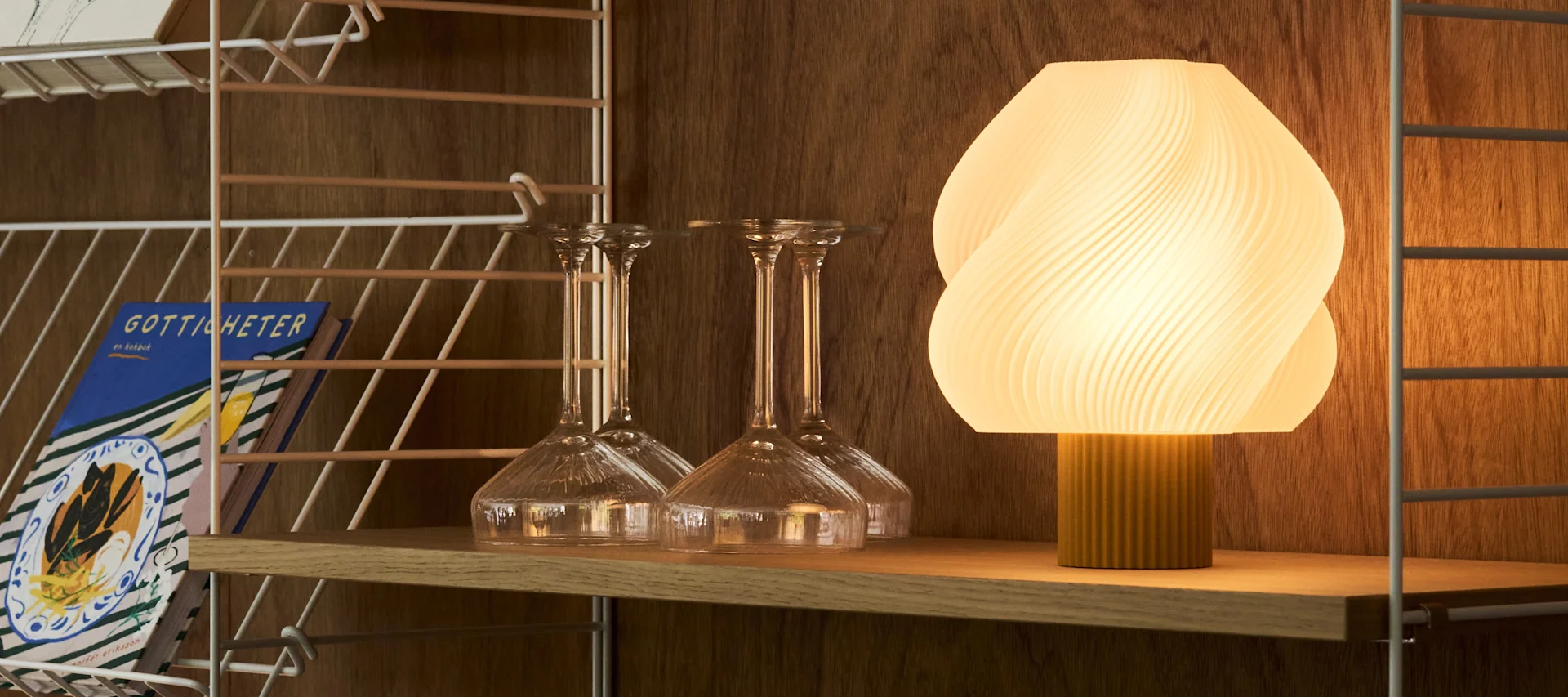 En upplyst, räfflad oval lampa och olika klara glas står på en träbarvagn mot en varm, texturerad vägg.