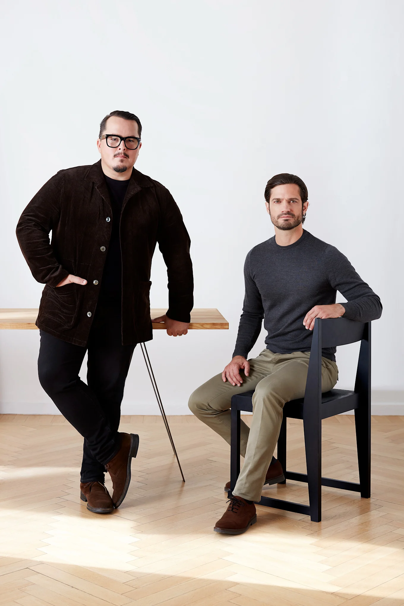 Två män, en står vid ett träbord i en brun manchesterjacka, den andra sitter på en svart stol i en grå skjorta.