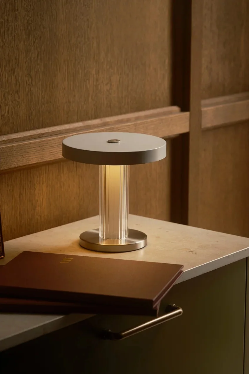En modern bordslampa med räfflad, transparent pelare och cirkulär bas och topp, som avger ett varmt sken på en bänkskiva bredvid ett brunt häfte.
