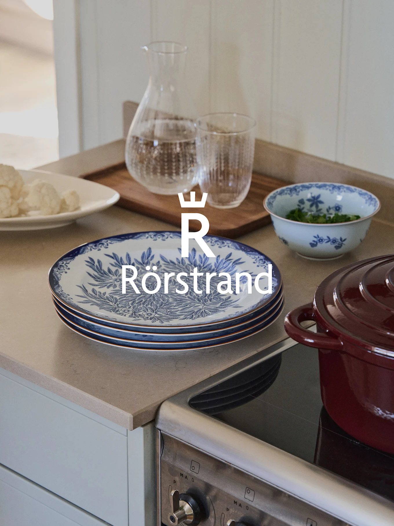 En köksbänk med Rörstrand-tallrikar, en karaff och en skål med blått blommönster, intill en röd gryta på en spishäll.