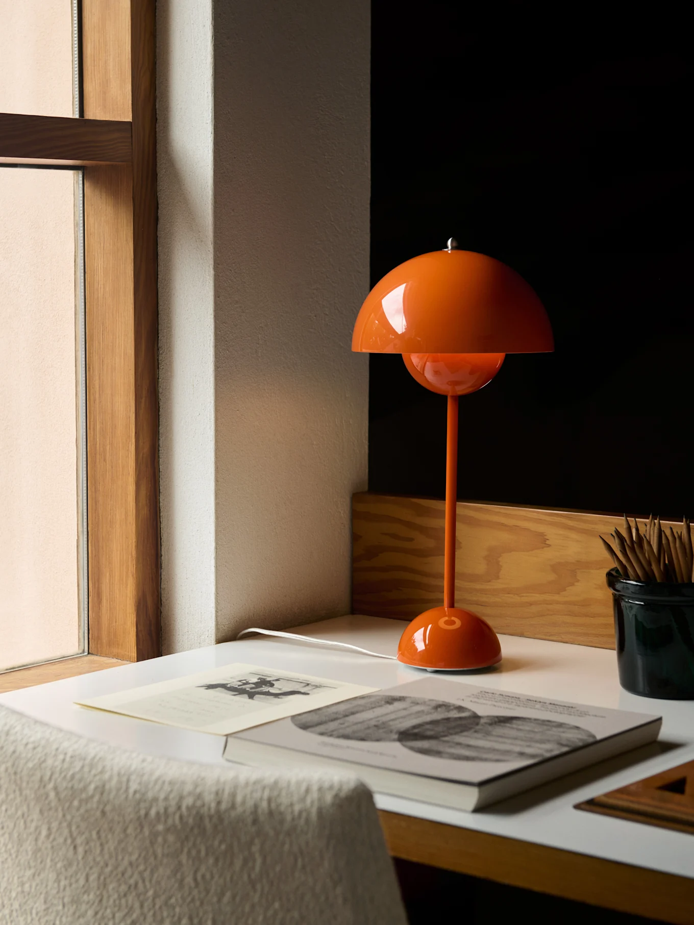 En orange svampformad bordslampa på ett vitt skrivbord bredvid ett fönster, med böcker och pennor.