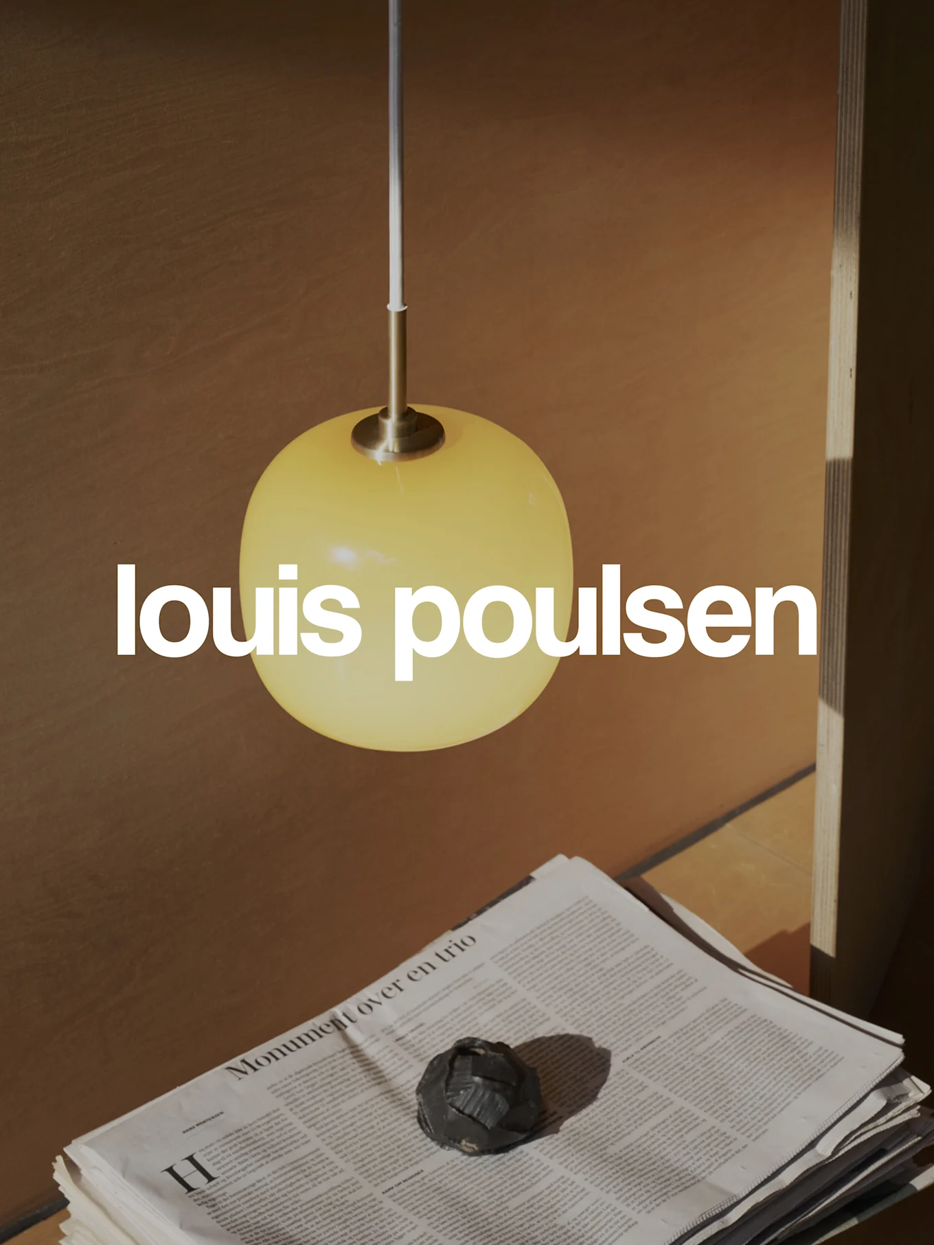 Louis Poulsen-logotyp över en varm scen med en gul sfärisk pendellampa, staplade tidningar och ett mörkt dekorativt föremål.
