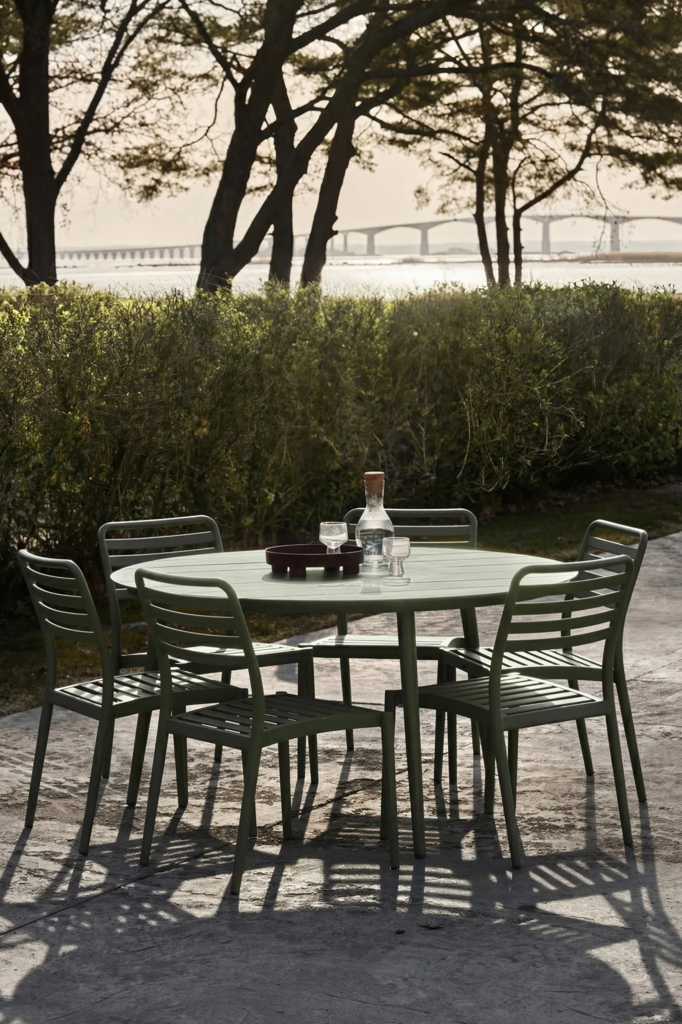 Grön utomhusmatgrupp med bord, fem stolar, vattenflaska och glas på uteplats. Bro över vatten i fjärran.