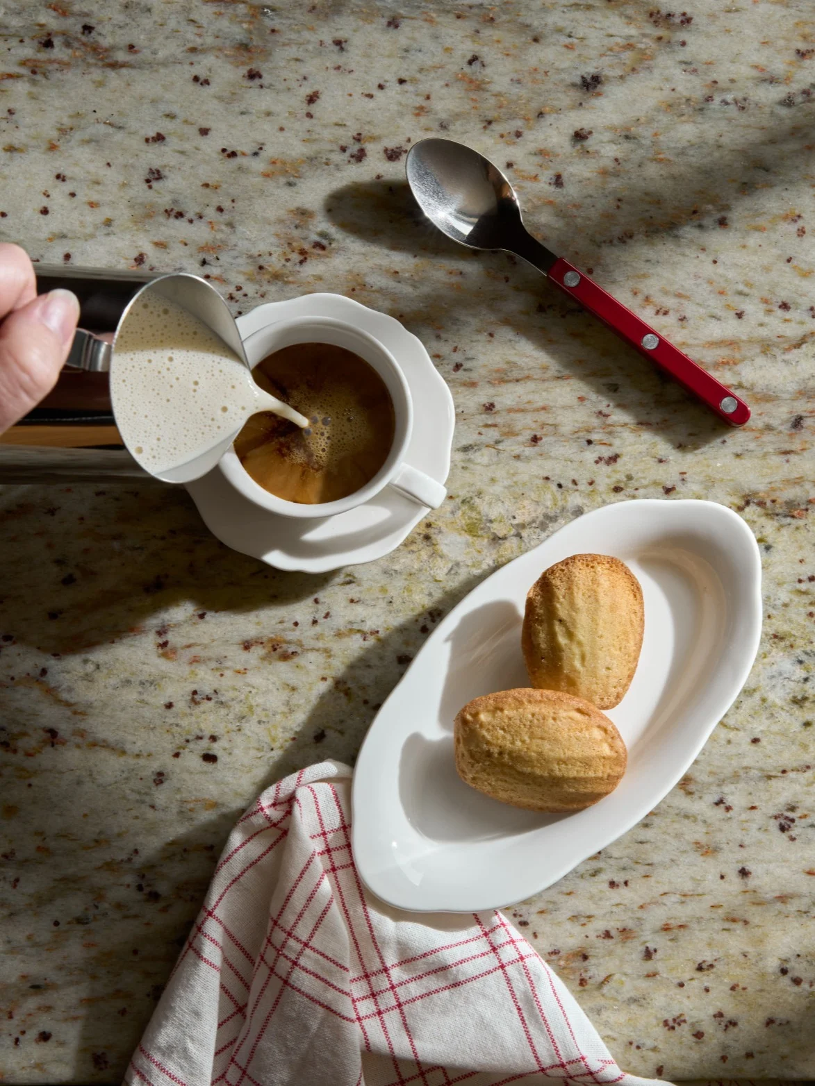 Hand häller skummad mjölk i kaffekopp bredvid madeleinekakor på en granitbänk.
