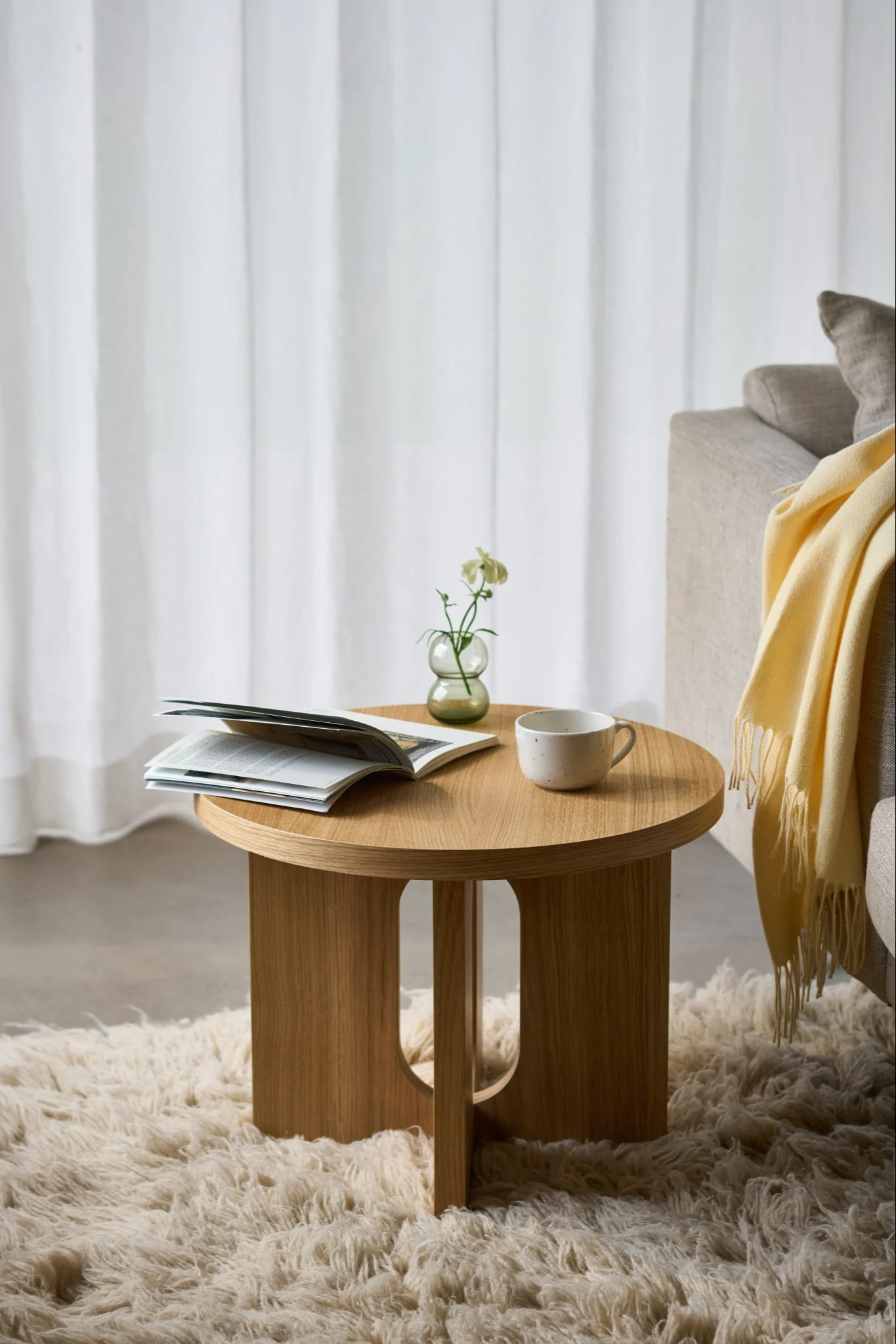 Runt sidobord i trä med en öppen bok, en mugg och en liten vas med en vit blomma på en fluffig matta, bredvid en soffa.