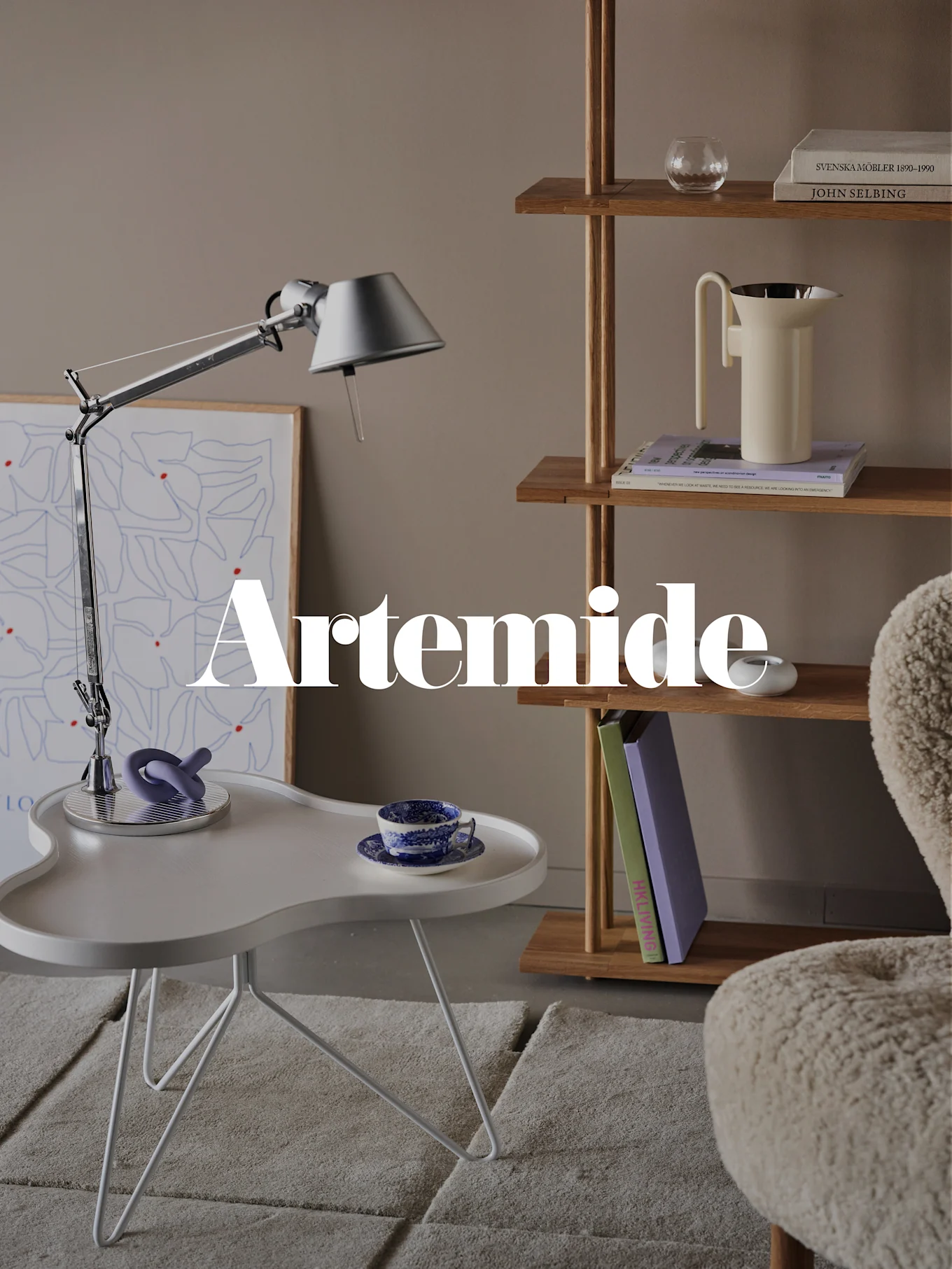 Ett modernt vardagsrum med en silverfärgad skrivbordslampa, vitt sidobord, träbokhylla och en krämfärgad fåtölj. Textöverlagring: 'Artemide'.