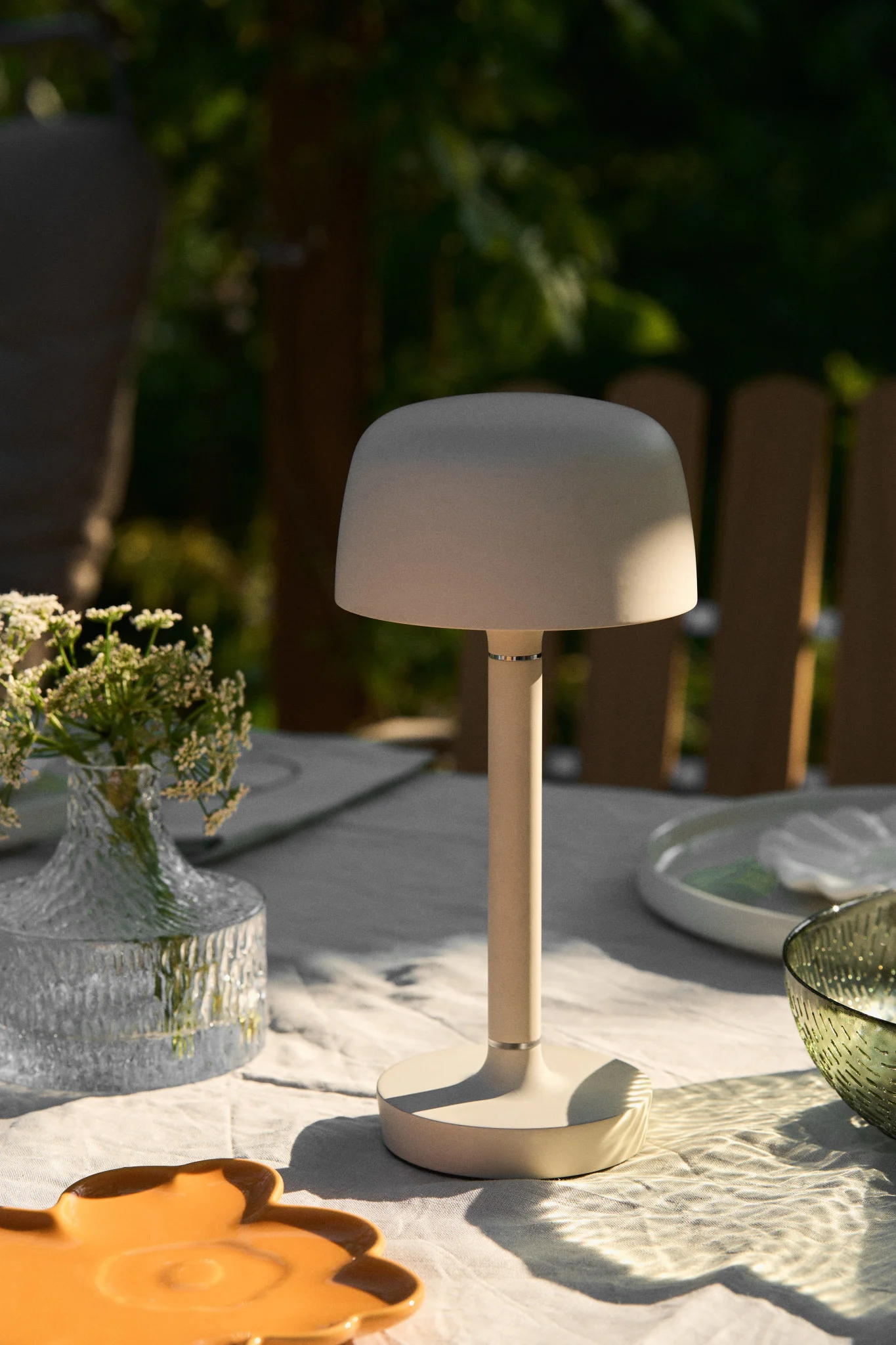 En svampformad beige bordslampa på ett vitt utomhusmatbord med blommor och porslin.