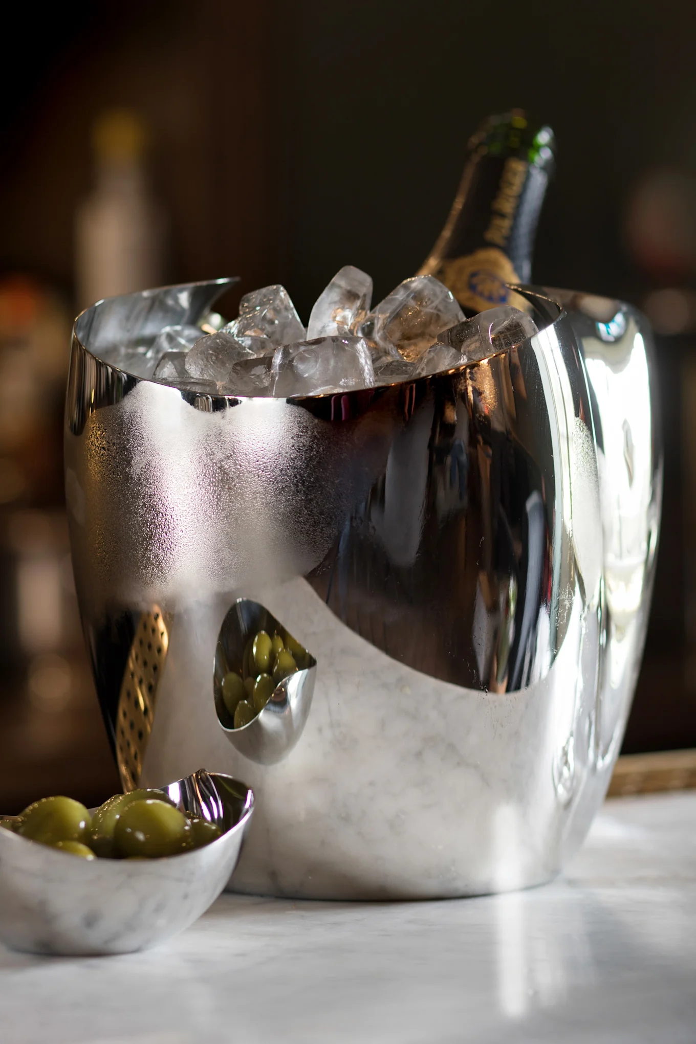 En reflekterande silverishink med en champagneflaska och is, bredvid två små skålar med gröna oliver på marmor.