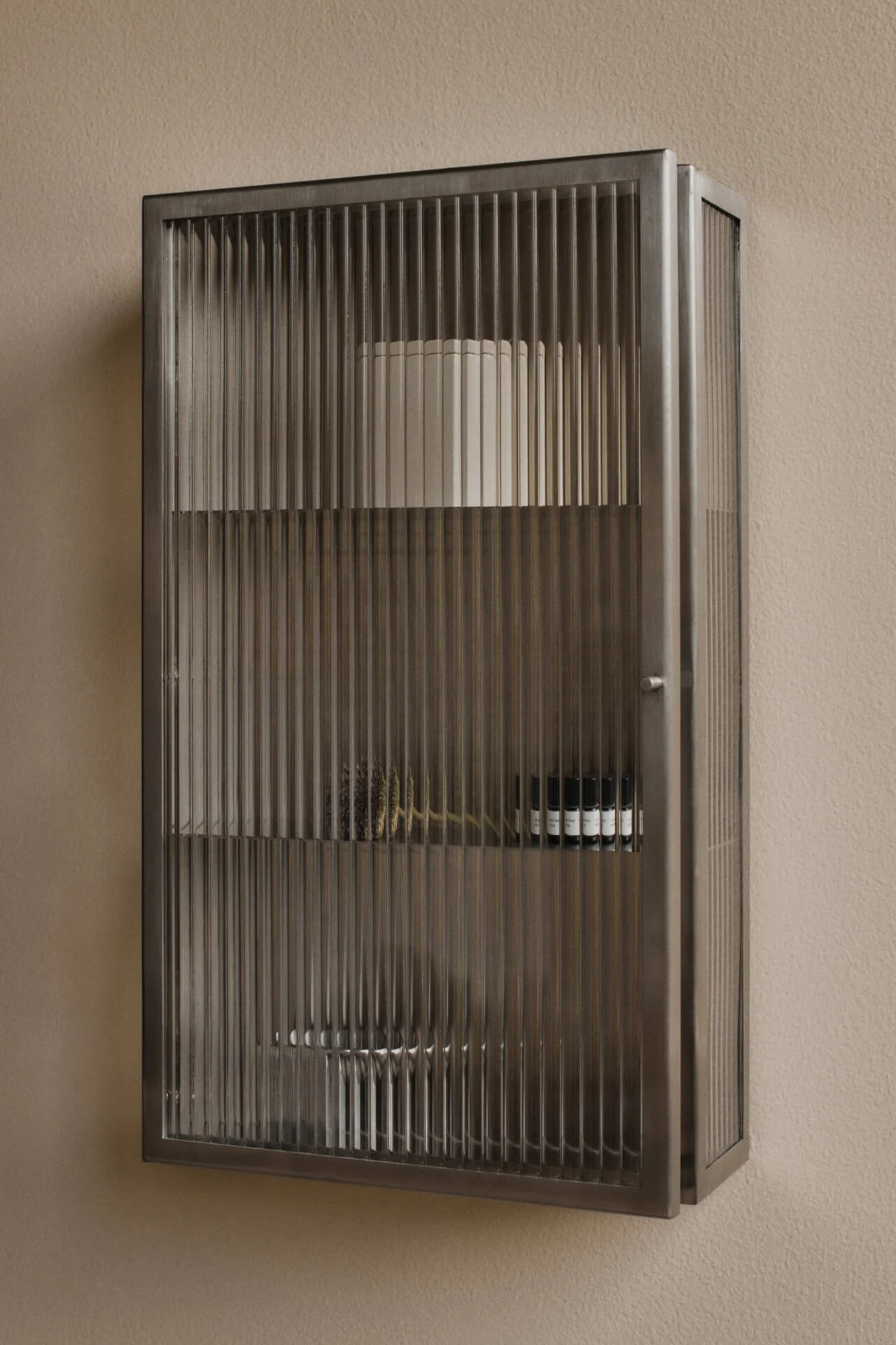Väggmonterat, mörkt metallskåp med räfflade glasdörrar på en beige vägg. Innehåller hyllor med en vit cylinder, torkade botaniska växter och små mörka flaskor.