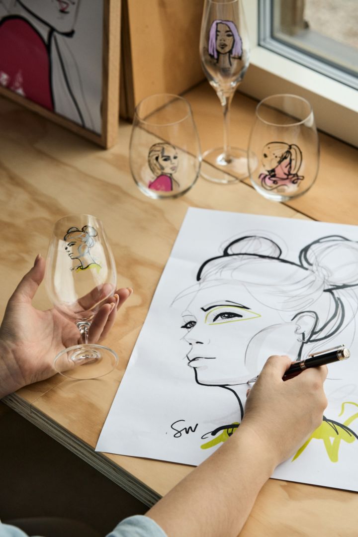 Kvinna håller i All about you vattenglas från Kosta Boda medan hon skissar en teckning på kvinnoansikte med den andra handen. 