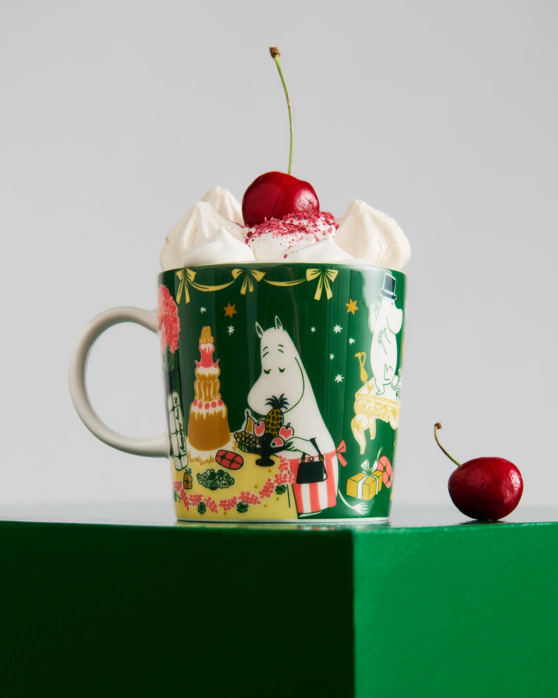 Mörkgrön mugg med Moomin-figurer, toppad med vispgrädde, rött strössel och ett körsbär. Ett annat körsbär ligger på den gröna ytan.