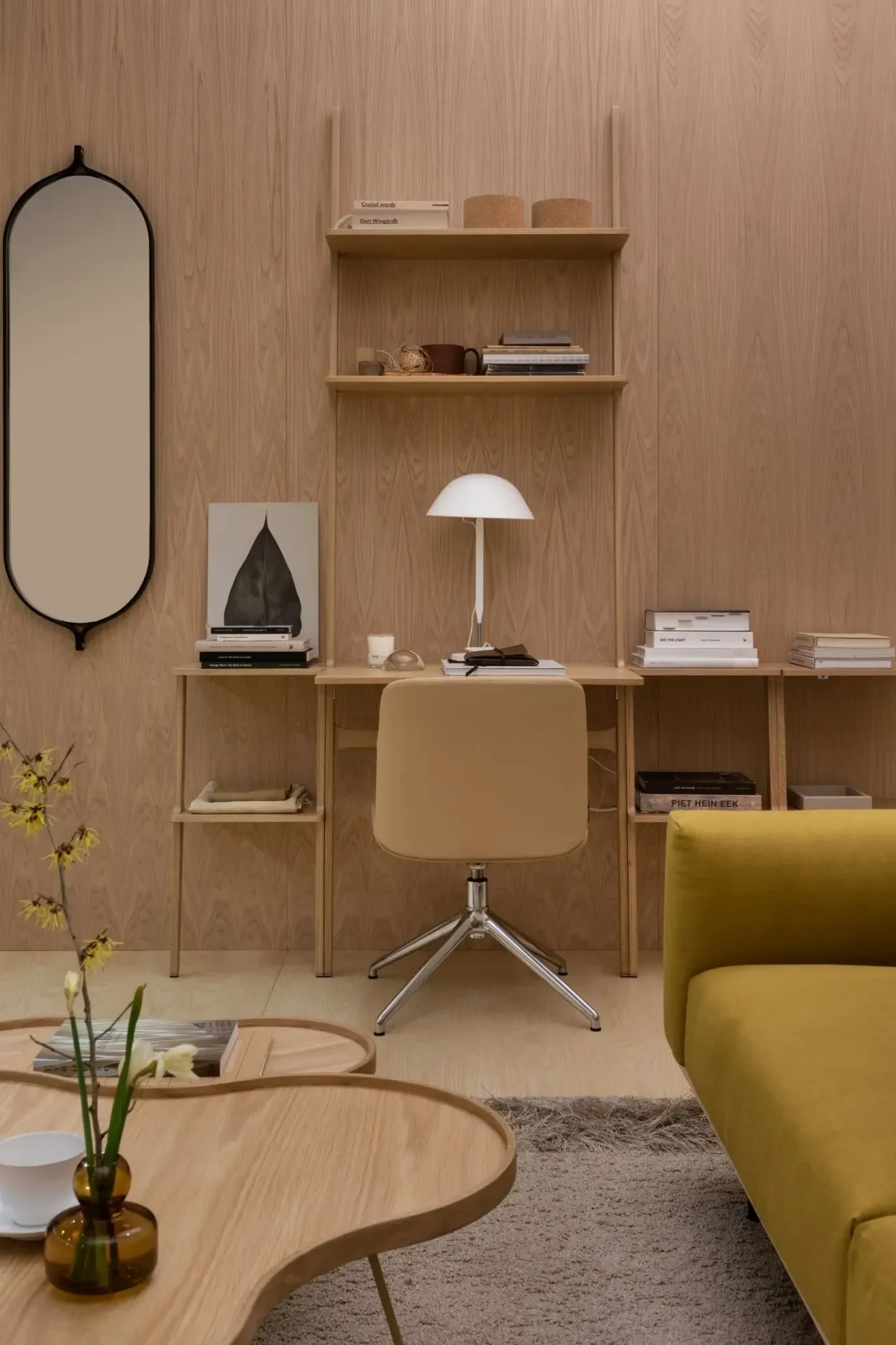 Ett minimalistiskt hemmakontor med ljus träpanel, en beige skrivbordsstol, vit lampa, hyllor, bredvid en gul soffa och soffbord.