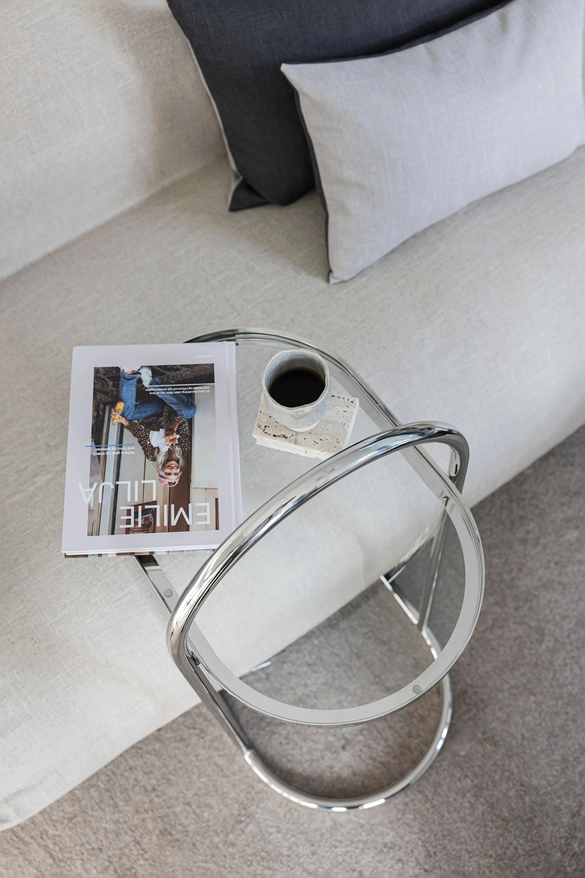 Översiktsvy av ett modernt vardagsrum med en ljus soffa, prydnadskuddar och ett sidoord i glas och krom med en tidning och kaffe.