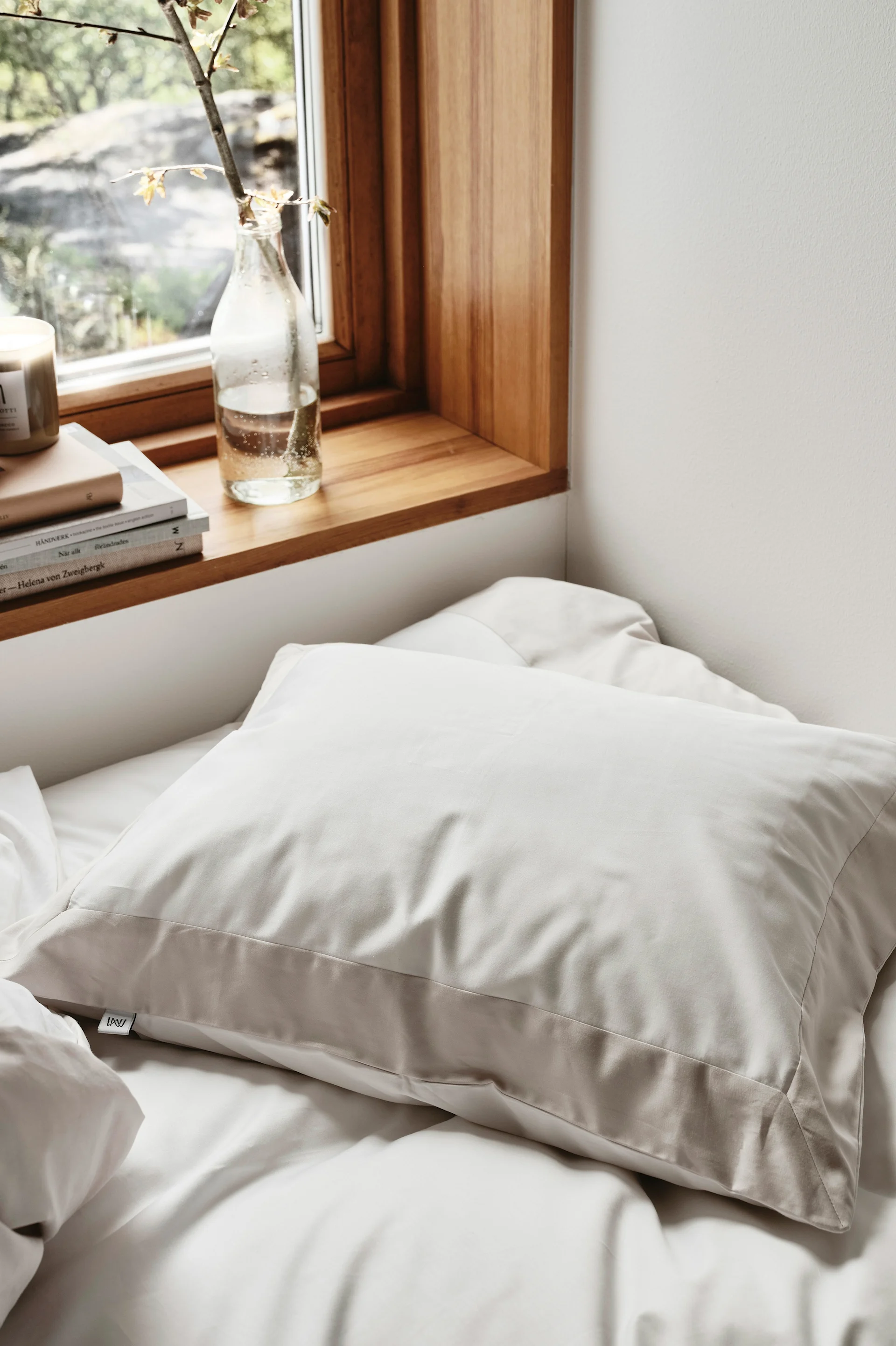 En ljusbeige kudde på en vit säng, bredvid ett fönster med en träfönsterbräda, böcker och en växtkvist i en flaska.