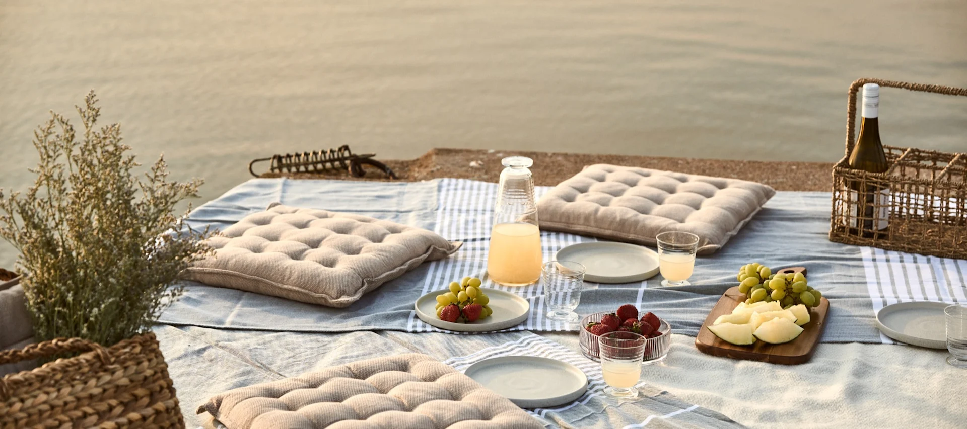 Picknick vid vattnet med kuddar, frukt och drycker på en filt.