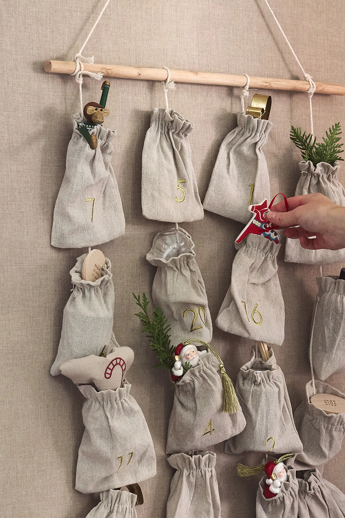 Tygadventskalender med numrerade påsar hängande från en träpinne; en hand placerar en röd Dalahäst.