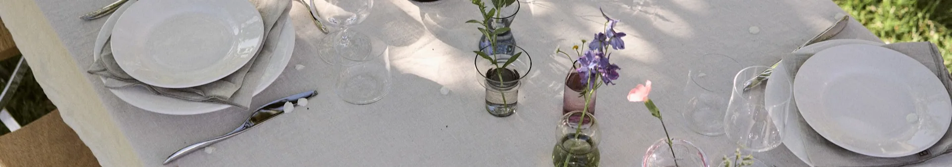 Utedukat bord med vita tallrikar, bestick och små vaser med lila, rosa och vita blommor på en ljus linneduk.