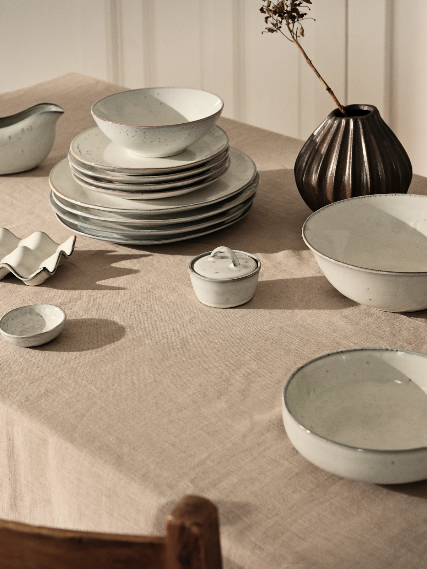 Ljusgrått keramiskt middagsset med staplade tallrikar och skålar på beige linneduk, med mörk vas och torkad gren.