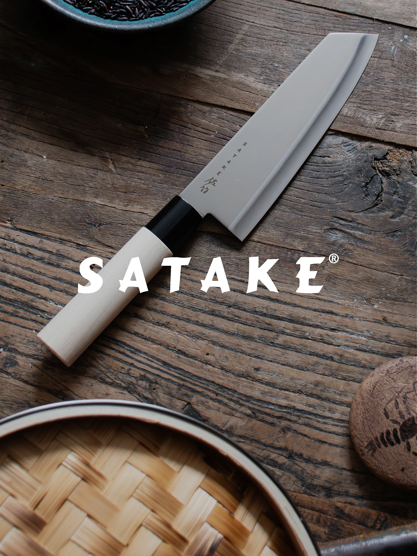 En Satake kockkniv med ljust handtag och grått blad på ett träbord, bredvid svart ris och en bambuångkokare.