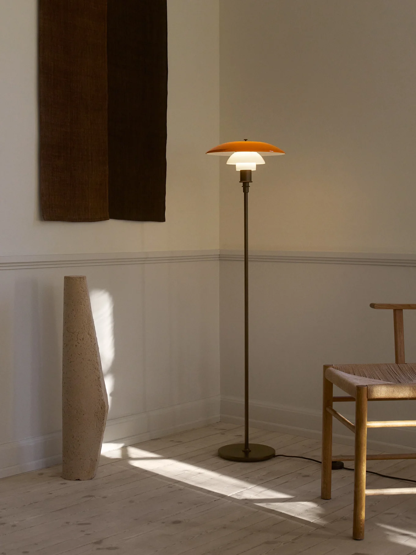 En modern, tänd golvlampa med en orangebrun skärm och vit diffusor, en hög texturerad vas och en trästol med flätad sits i ett soligt rum.