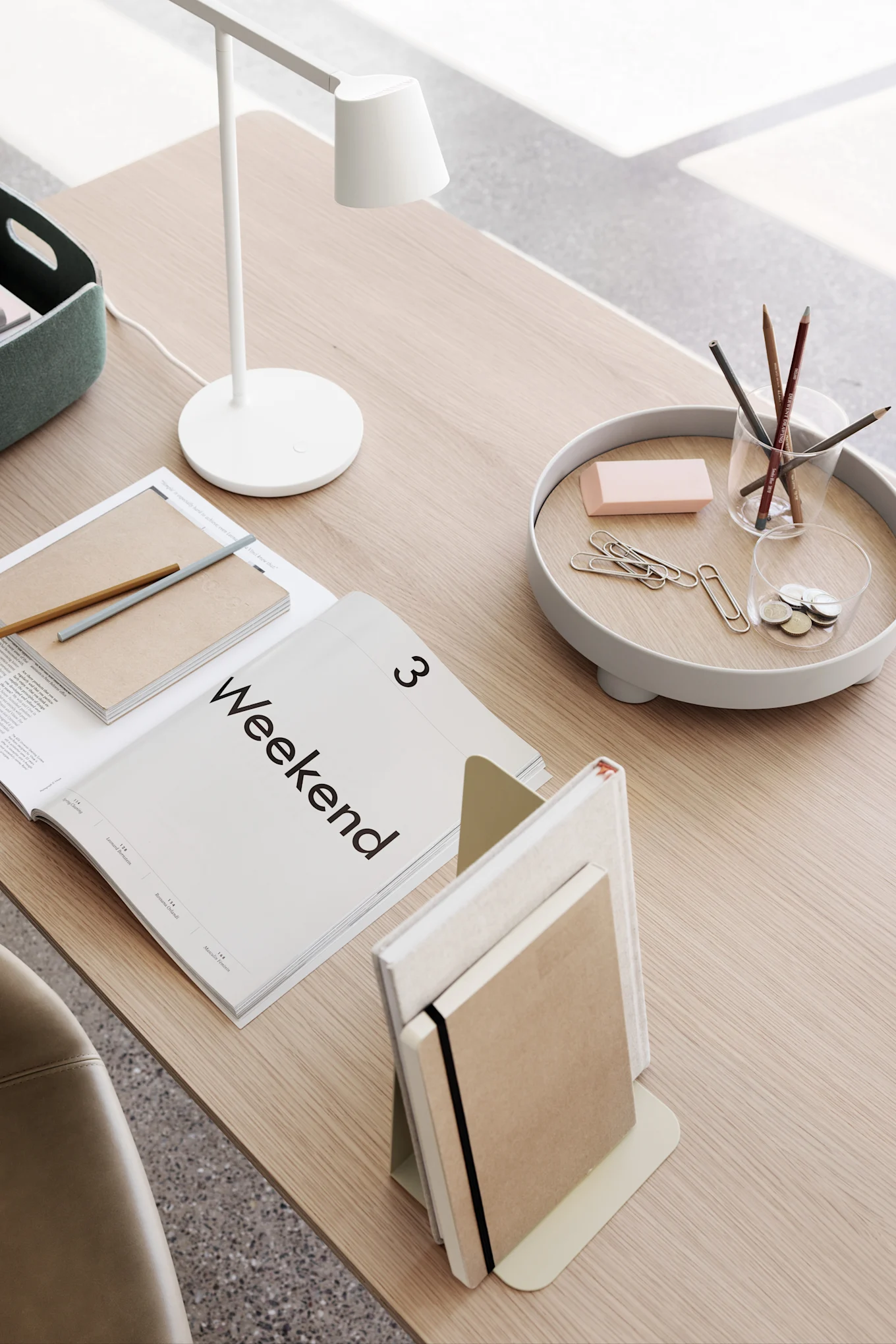 Minimalistisk skrivbordsuppsättning med vit lampa, öppen 'Weekend'-tidning, anteckningsböcker och skrivbordsaccessoarer på en bricka.