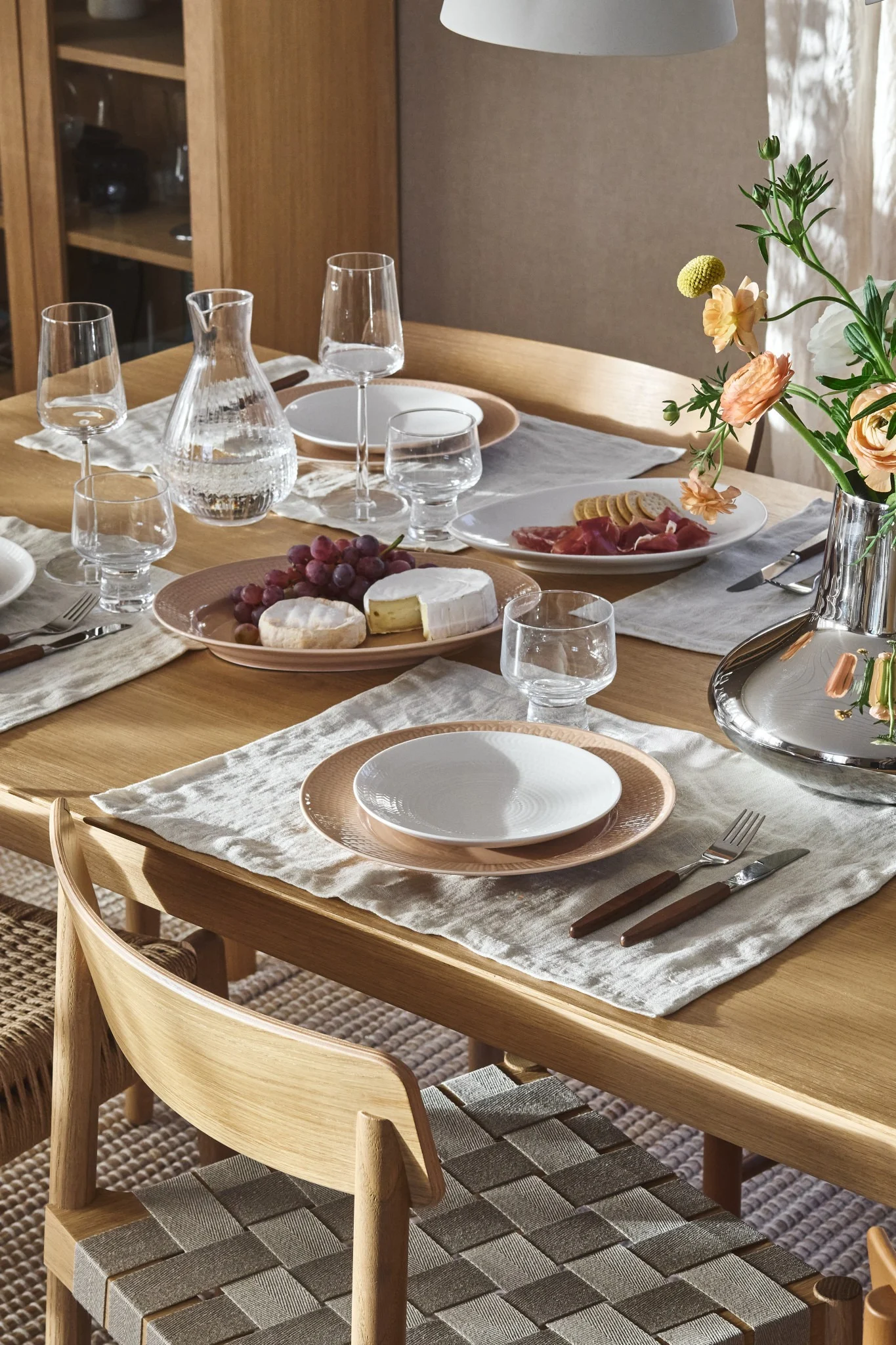 Matsalsbord med tallrikar, glas, karaff, vindruvor, ostar, charkuterier och blommor. Flätade stolar. Porslinet kommer ifrån Rörstrand Jubilee kollektion. 