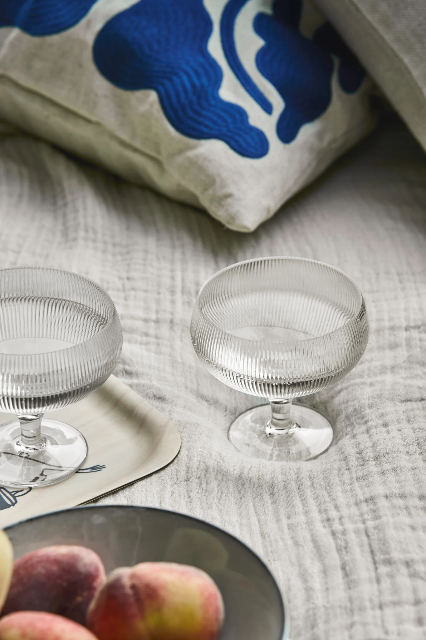 Två räfflade coupeglas i glas på ljust grått tyg, med persikor i en skål och en blåmönstrad kudde.